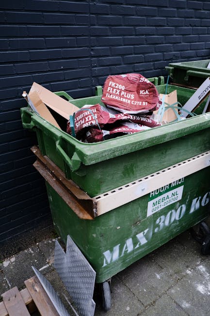 A green wheeled waste container filled with black and red bagged rubbish, placed on a paved outdoor area against a dark blue brick wall. The bags appear to contain construction or DIY waste, featuring printed labels with text such as 'FK200 FLEXY PLUS.' The container is partially obscured by a wooden slatted platform positioned underneath, and a metal ramp or object leans against its side. Nearby, a second similar container is visible, also filled with waste bags, indicating an area used for private rubbish storage or collection outside a building. The environmental context suggests a residential or commercial property where waste is temporarily stored before collection, highlighting issues related to access and the need for proper waste management services, such as those offered by Waste Disposal Haringey, which specializes in rubbish and waste removal solutions. The scene is lit by natural daylight, giving a clear view of the textures of the plastic bags, the metal wheels of the container, and the brickwork behind. This setup may relate to alternative waste handling methods where external storage is necessary due to access restrictions or collection limitations.