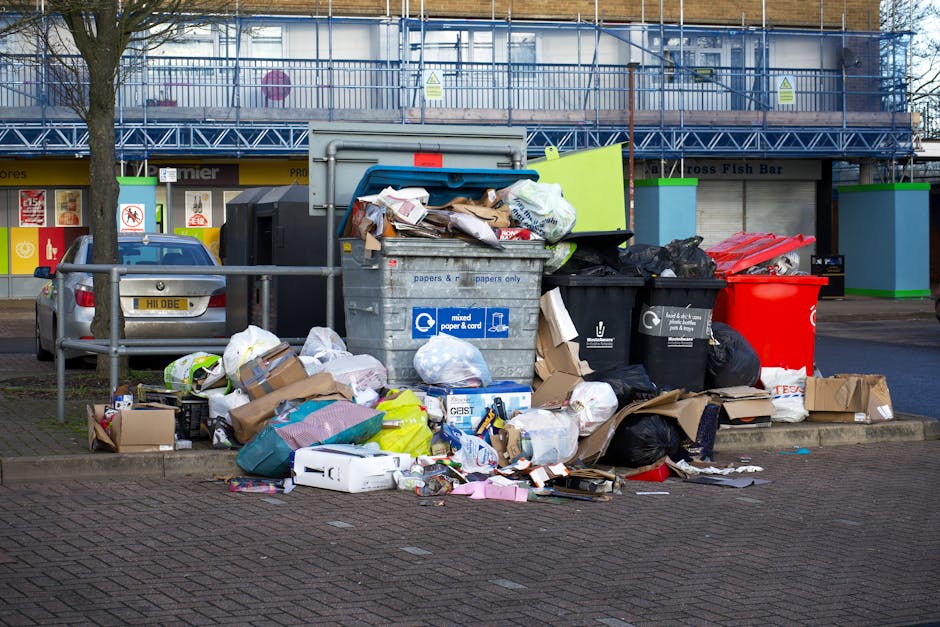 A green wheeled waste container filled with black and red bagged rubbish, placed on a paved outdoor area against a dark blue brick wall. The bags appear to contain construction or DIY waste, featuring printed labels with text such as 'FK200 FLEXY PLUS.' The container is partially obscured by a wooden slatted platform positioned underneath, and a metal ramp or object leans against its side. Nearby, a second similar container is visible, also filled with waste bags, indicating an area used for private rubbish storage or collection outside a building. The environmental context suggests a residential or commercial property where waste is temporarily stored before collection, highlighting issues related to access and the need for proper waste management services, such as those offered by Waste Disposal Haringey, which specializes in rubbish and waste removal solutions. The scene is lit by natural daylight, giving a clear view of the textures of the plastic bags, the metal wheels of the container, and the brickwork behind. This setup may relate to alternative waste handling methods where external storage is necessary due to access restrictions or collection limitations.