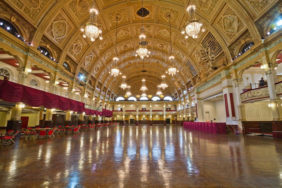The image displays a large, ornate indoor hall with high, vaulted ceilings decorated with intricate patterns and multiple elegant chandeliers hanging from the ceiling. The polished wooden floor reflects the warm lighting, and the space is mostly empty except for several rows of red chairs arranged along one side. There are decorative drapes in a deep red colour hanging from the upper balcony area, which features a railing with detailed mouldings. The walls are painted in light hues with cream and gold accents, and arched windows allow natural light to diffuse into the spacious interior. The setting appears to be a historical or formal event venue, with a spacious layout suitable for gatherings or functions. The scene is well-lit with a warm, welcoming atmosphere, fitting for an alternative event space or venue that might host private functions instead of traditional public uses, indirectly relating to the context of waste management and venue cleaning services provided by Waste Disposal Haringey for event spaces like this.