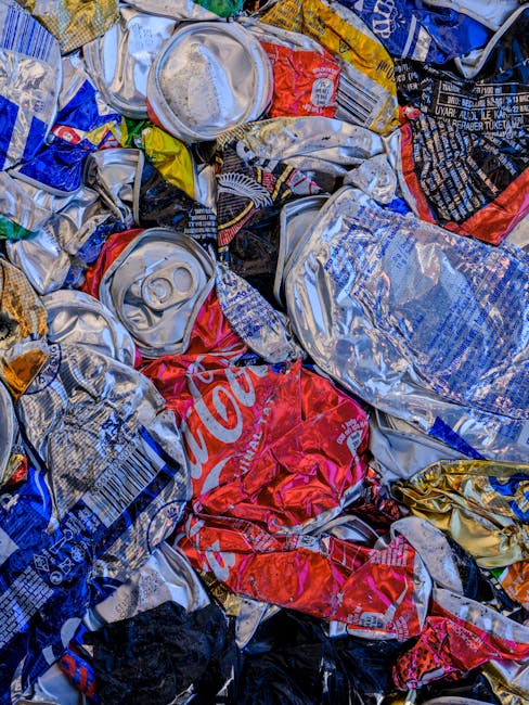 A close-up image of a pile of crushed aluminium cans and plastic beverage bottles, predominantly silver, red, blue, and yellow in color, with some cans showing brand labels such as Coca-Cola. The cans and bottles are jumbled together, with some overlapping and others partially flattened or dented. The surface textures include smooth, reflective metal and glossy plastic, with print and label details visible despite compression. The background consists of additional discarded containers, suggesting a collection of recyclable waste, likely in the context of local rubbish removal or recycling services. The scene is lit with natural or neutral lighting, emphasizing the metallic and plastic surfaces, indicative of waste materials ready for disposal or transfer to a recycling facility, possibly handled by companies like Waste Disposal Haringey, focusing on environmentally responsible waste management and alternative collection methods.