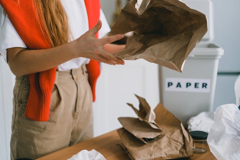 A person wearing a beige skirt, a white shirt, and an orange sleeveless cardigan is seen placing crumpled brown paper bags into a recycling bin designated for paper, which has a label reading 'PAPER' on its front. The scene appears to be set indoors, possibly in an office or kitchen environment, with a light-colored wall in the background. On the wooden table surface nearby are additional crumpled paper bags, a white plastic bag, and a small glass jar, indicating waste sorting or preparation for rubbish removal. The recycling bin is positioned on the right side of the image, and the individual’s hands are in the process of discarding or organizing the paper waste. This scene illustrates household or office waste disposal practices, aligned with guidelines for proper rubbish management, and reflects actions consistent with alternative waste handling solutions facilitated by waste disposal services such as Waste Disposal Haringey.