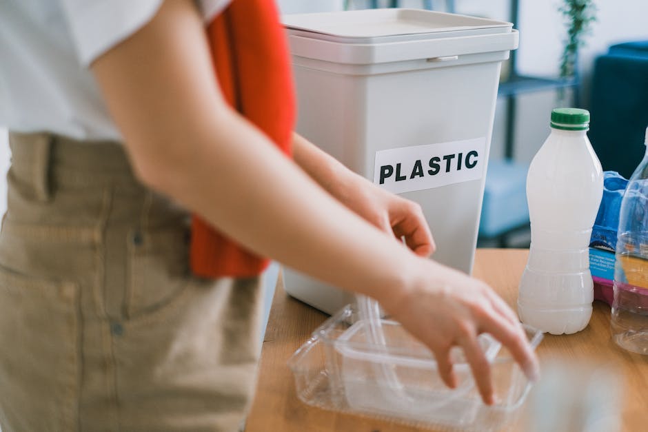 In the foreground, a person’s torso and arms are visible as they handle a clear plastic container, positioning it on a wooden surface. The individual is wearing beige trousers and a white top, with a red cloth or item tucked under their arm. Behind them, a white recycling bin with a black label reading 'PLASTIC' is prominently placed on the table, indicating a designated area for plastic waste collection. To the right of the bin, there are several plastic bottles, including a transparent bottle with a green cap and other assorted bottles, some of which are partially filled or capped. The background features an indoor environment with a soft, neutral lighting and hints of blue and green elements, possibly furniture or decor. The scene conveys a moment of sorting or preparing household waste for disposal or recycling, aligning with waste management practices, and supported by the presence of waste collection containers as used by companies like Waste Disposal Haringey for rubbish removal or alternative waste handling outside of council services.