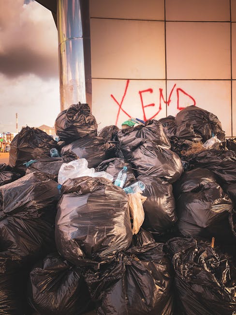 A large pile of black garbage bags filled with waste is stacked against a modern building with a tiled exterior. Several clear plastic water bottles and small plastic items are visible spilling from some of the bags. Behind the pile, on the wall, the word 'XEND' is spray-painted in red graffiti. The scene is outdoors, and the lighting suggests late afternoon or early evening, casting a warm glow over the area. The environment appears to be an urban setting, possibly a service alley or designated disposal zone, highlighting a typical example of unmanaged waste accumulation that could be subject to private rubbish collection services such as those offered by Waste Disposal Haringey, which specializes in alternative waste handling solutions to avoid issues like illegal dumping or on-site clearance problems.