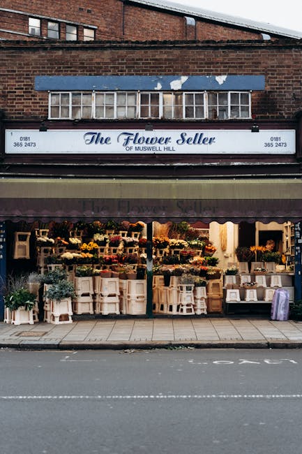 The exterior of a flower shop named 'The Flower Seller of Muswell Hill' features a large white sign with blue lettering, positioned above a dark maroon awning. The shopfront displays an array of flower arrangements, potted plants, and bouquets, arranged on tables and shelves that extend across the storefront. The pavement outside is paved with rectangular concrete slabs, and the street in front has visible lane markings. The shop is set within a brick building with a second-story window and a small vent or skylight above. The scene appears during daytime, with natural light illuminating the display of flowers and plants, highlighting their vibrant colors and textures. The overall setting suggests a typical small town or suburban commercial area, where independent businesses like this flower shop might independently handle waste and disposal, potentially involving rubbish removal services to manage waste from daily stock replenishment or clearance of plant debris. Waste Disposal Haringey’s presence in the local waste management sector can be inferred as one of the options for such businesses seeking reliable rubbish collection and vegetation waste disposal solutions.