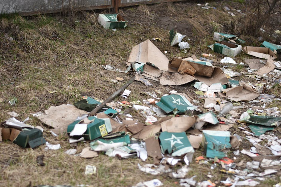 A collection of mixed waste materials piled on the ground in an outdoor area, including black plastic rubbish bags, a yellow plastic container, and an old, dirty, cream-colored upholstered chair with visible stains and wear. The debris is situated in front of a low stone wall composed of irregularly shaped, rough-textured stones. In the background, there are green trees and shrubbery, a tall metal pole with electrical wires overhead, and a partially visible large structure with a curved roof, likely part of a sports or community facility. The scene is illuminated by natural daylight, casting shadows on the gravel surface beneath the waste, which suggests an unauthorised rubbish dump or an on-site clearance situation handled by a private waste disposal company such as Waste Disposal Haringey, highlighting the need for professional rubbish removal services to manage such waste accumulation effectively.
