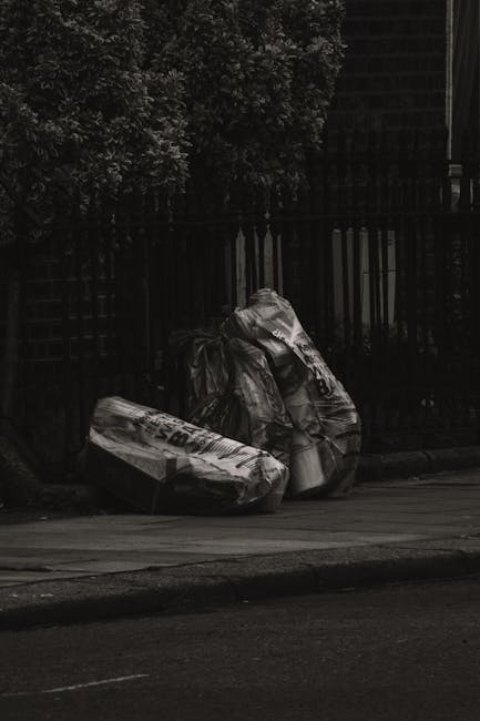 Three large black plastic rubbish bags, filled and tied at the top, are placed on a paved sidewalk next to a black metal fence with vertical bars. The bags appear to contain household waste or similar debris, with some loose material visible at the edges. Behind the fence, dense foliage of trees or bushes is visible, contributing to a shaded environment. The scene is illuminated by diffuse natural light, creating subtle shadows on the pavement and bags. The setting suggests an urban or residential area where private rubbish disposal, such as bagged waste outside a property, is evident. The presence of Waste Disposal Haringey’s mention implies the scene relates to alternative waste handling or rubbish removal services, where waste is temporarily stored for collection or disposal outside the property boundary. The overall composition emphasizes the practical aspect of on-site clearance tasks often undertaken by professional rubbish removal services to manage household waste effectively, particularly in urban environments requiring prompt and organized disposal methods.