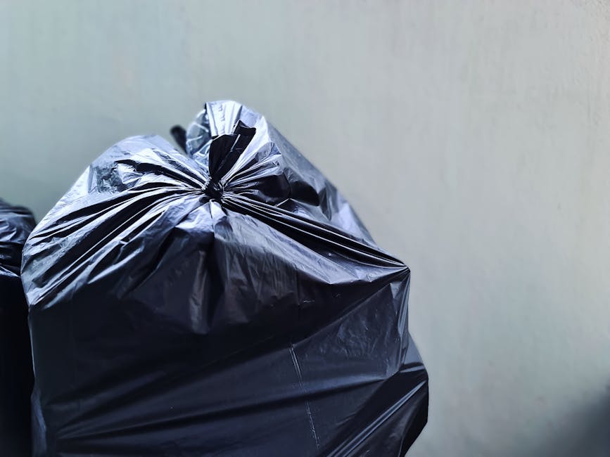 Close-up of several black plastic rubbish bags filled with waste, tied securely at the top and placed against a plain, neutral-colored wall. The bags have a glossy, slightly crinkled texture, indicating they are made of standard polyethylene material. They are positioned on the ground, with one bag prominently in the foreground and others stacked or leaning slightly behind it. The scene appears to be in an indoor or sheltered area, possibly a driveway or storage space used for waste collection before removal. The lighting is even, with minimal shadows, suggesting a well-lit environment. Waste Disposal Haringey may manage similar on-site clearance or private waste handling services, as these bags are prepared for collection outside the property, reflecting the kind of alternative rubbish removal some clients opt for instead of local authority collection.