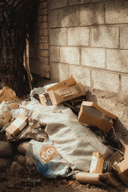A pile of mixed waste materials stacked against a rough-textured concrete brick wall, including cardboard boxes of various sizes, some flattened or partially torn, and large woven polypropylene sacks filled with rubbish. The cardboard boxes are light brown, with some showing printed labels and barcode markings, and are positioned on top of and around the sacks. The sacks are off-white with visible fabric weave patterns and branding in orange and blue. The waste materials are situated on uneven ground with small rocks and dirt, adjacent to a dark, textured tree trunk on the left side of the image. The scene is outdoors, with natural lighting illuminating the debris and surrounding environment, indicating an informal or non-commercial waste disposal area, which Waste Disposal Haringey might be involved in managing as part of private or on-site rubbish clearance services.