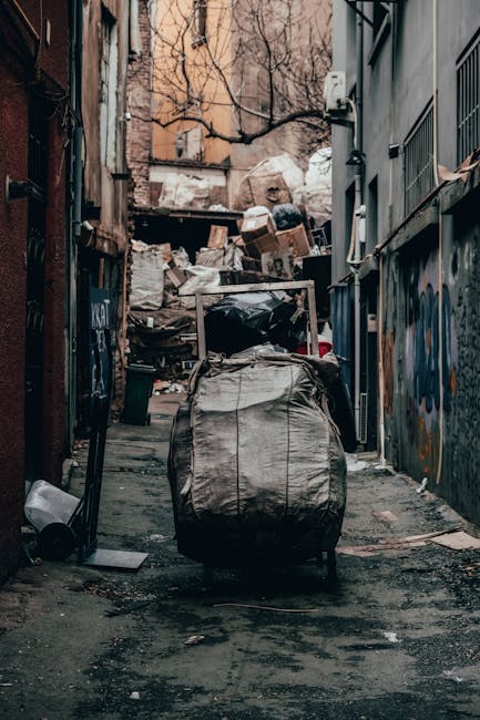 A narrow urban alleyway cluttered with accumulated rubbish and debris, with a large black trash bag and a wheeled waste container in the foreground. Behind these, there are overflowing piles of flattened cardboard boxes, plastic bags, and miscellaneous waste materials stacked against a weathered brick wall and a corrugated metal fence. The surrounding environment includes the facades of closely spaced buildings, one with a mural or graffiti on the right wall, and another with exposed brickwork and some graffiti on the left. The alley appears to be used for private waste disposal rather than official refuse collection, with a mix of discarded packaging and general waste materials. The lighting is natural, suggesting daytime with overcast skies, highlighting the textures of the garbage and the urban decay of the area, which aligns with independent rubbish clearance activities often handled by local waste disposal services like Waste Disposal Haringey.