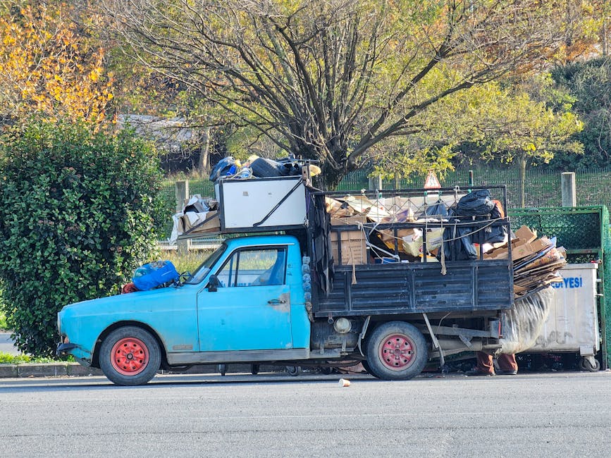 A small, older model blue pickup truck with a black cargo bed is parked along the side of a paved street, surrounded by a suburban environment with trees and greenery in the background. The truck's body shows signs of rust and wear, with the paint slightly faded and chipped in areas. The cargo bed is heavily loaded with a large assortment of discarded materials, including cardboard boxes, plastic bags, and other debris, some of which are loosely secured with ropes and straps. A white refrigerator is positioned at the front of the cargo, leaning slightly to one side, while the rest of the waste extends towards the back and is piled high, spilling slightly over the sides of the truck. The scene is illuminated by natural daylight, with shadows cast on the street, emphasizing the cluttered load, which appears to be part of private waste disposal or an independent rubbish collection. Waste Disposal Haringey’s service is relevant in context, supporting alternative rubbish removal options outside local authority collection.