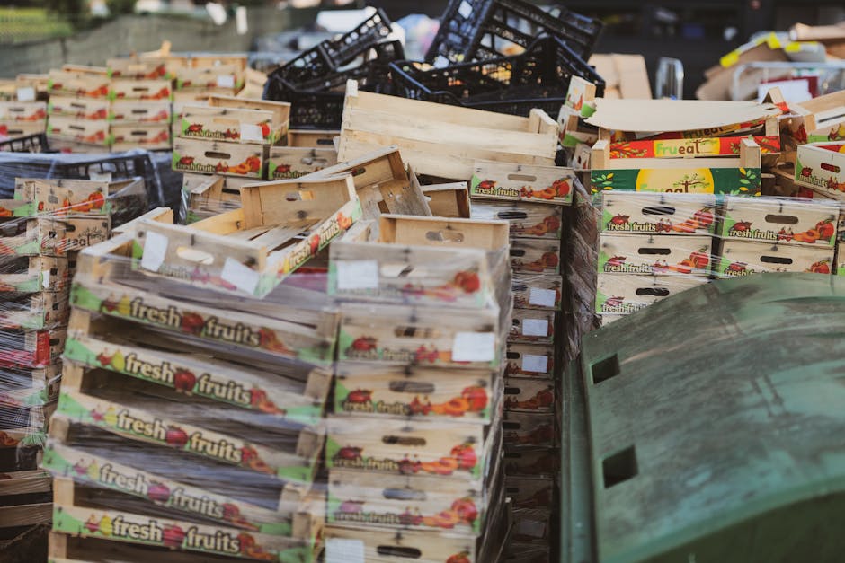 A large outdoor area filled with numerous stacks of wooden fruit boxes, featuring colorful branded labels with illustrations of fresh vegetables and the text 'fresh fruits.' The boxes are stacked in various orientations, some leaning against each other and others piled unevenly. Behind the boxes, there are dark plastic crates and additional cardboard packaging visible. The scene appears to be part of a waste collection or clearance area for discarded produce containers, indicative of an alternative waste handling process or private disposal service. The environment is outdoors, with a hint of natural light illuminating the scene, and the overall visual emphasizes bulk waste of wooden packaging commonly associated with fruit or vegetable transport. Waste Disposal Haringey’s presence in this context suggests a professional service for managing such waste streams, possibly during on-site clearance or bulk pick-up activities. The arrangement and materials reflect typical waste management procedures for packaging waste in a commercial or wholesale setting, supporting the local rubbish removal options available in the area.